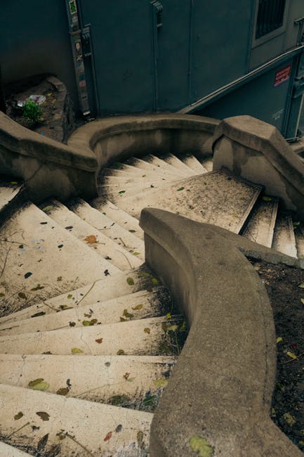 A set of outdoor stone stairs with curved handrails on each side, descending from an upper level to a lower pavement area. The steps are made of light-colored stone with visible dirt and fallen leaves