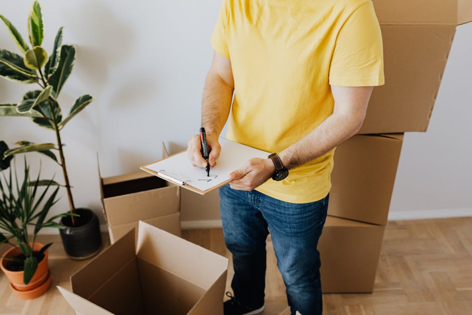 A man wearing a yellow t-shirt and jeans is standing indoors, surrounded by several large cardboard moving boxes. He is holding a clipboard in his left hand and using a black marker with his right han