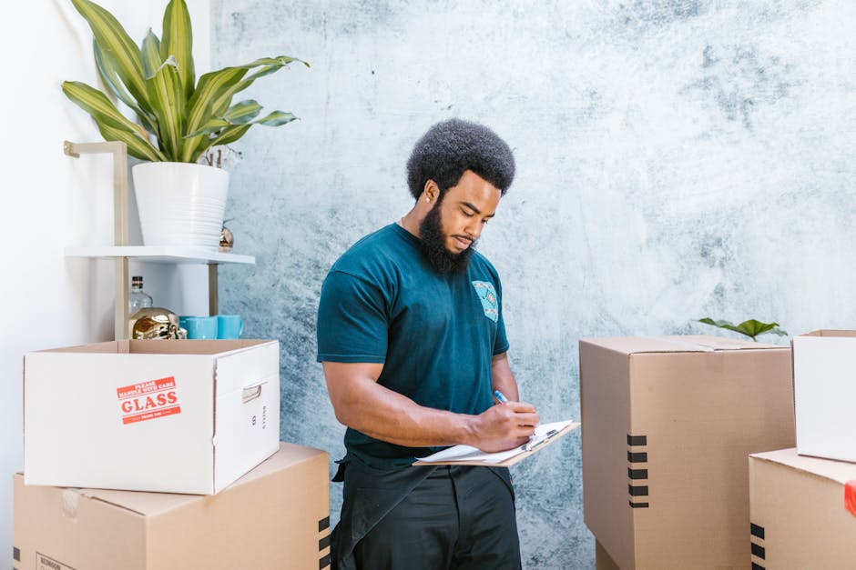 A man wearing a yellow t-shirt and jeans is standing indoors, surrounded by several large cardboard moving boxes. He is holding a clipboard in his left hand and using a black marker with his right han
