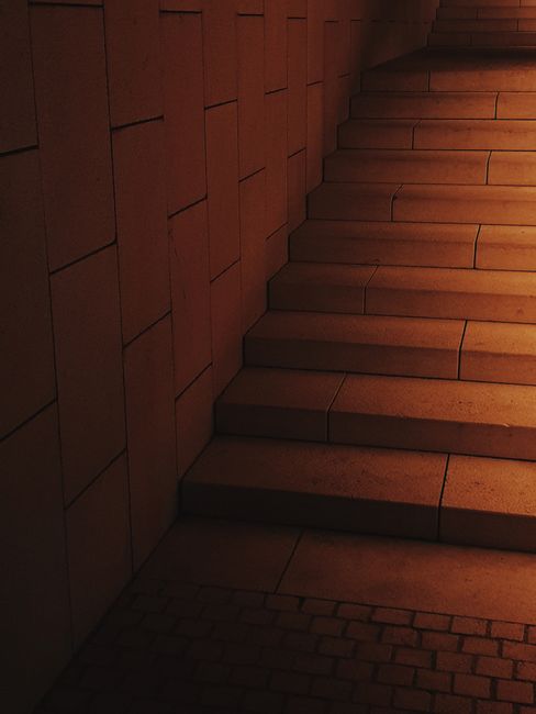 An indoor scene showing a section of a wooden staircase with sunlight casting shadows onto the treads. To the left, part of a patterned tile floor with a geometric design in shades of beige, brown, an