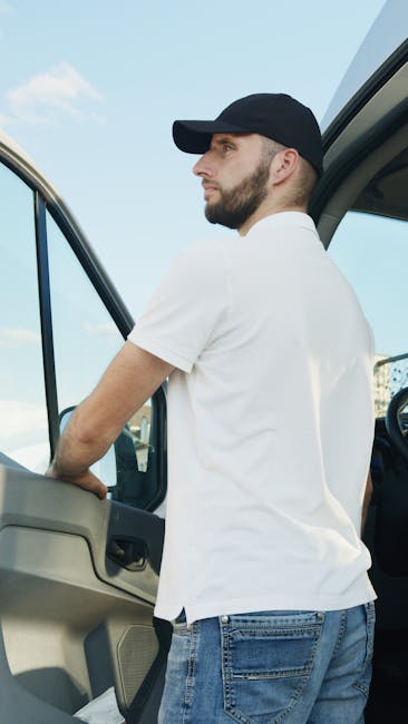 A man wearing a grey cap, glasses, a beige and grey jacket, black gloves, and dark pants, is opening the sliding side door of a white cargo van positioned on a residential driveway. The van has black